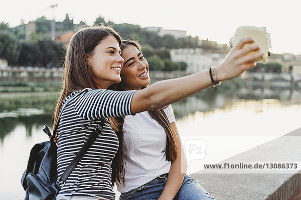 Freunde fotografieren sich selbst mit Sofortbildkamera  während sie auf einer Stützmauer am Fluss Arno in der Stadt sitzen