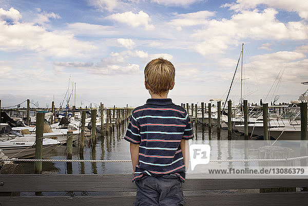 Rear view of boy standing on bridge by harbor against cloudy sky