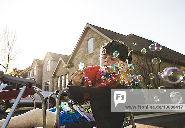 Boy with fractured leg blowing bubbles while sitting on chair against house during sunset