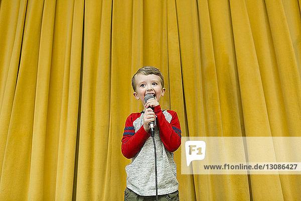 Selbstbewusster Junge singt am Mikrofon auf der Bühne gegen gelbe Vorhänge