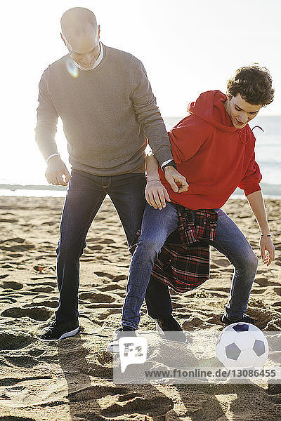 Full length of father and son playing soccer at beach during sunny day