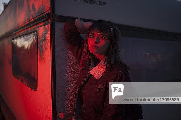 Portrait of confident young woman wearing jacket while standing by wall in red illuminated room