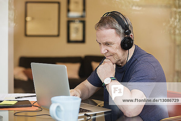 Senior man listening music while using laptop seen through glass window