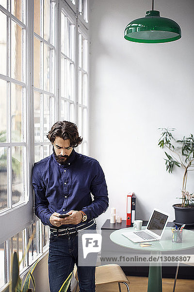 Businessman using mobile phone while leaning on window in office