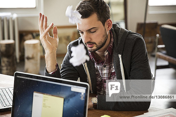 frustrated worker sitting by laptop on table with crumbled paper in mid-air at office