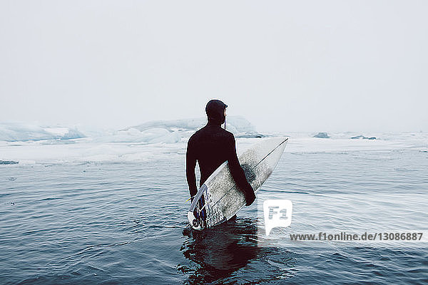 Mann trägt Surfbrett  während er im Winter im eisigen Meer vor klarem Himmel steht