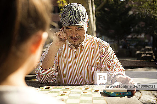 Nachdenklicher älterer Mann spielt Damespiel mit Enkelin am Tisch im Park