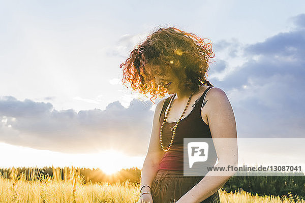 Lächelnde Frau schaut nach unten  während sie bei Sonnenuntergang auf dem Bauernhof vor bewölktem Himmel steht