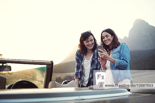 Lächelnde Frau zeigt Freundin bei Sonnenuntergang ihr Handy im Cabrio