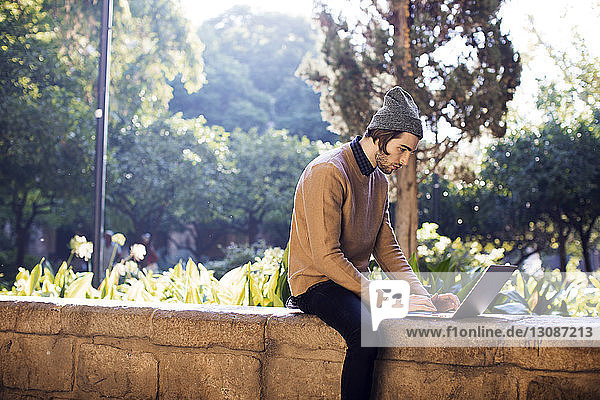 Side view of man using laptop computer while sitting on retaining wall