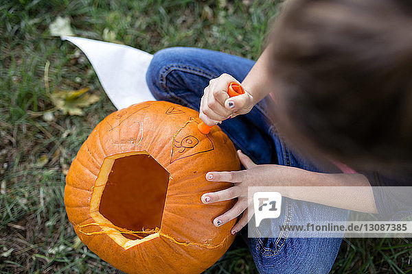 Hochwinkelaufnahme eines Mädchens  das an Halloween auf einem Feld im Hof sitzt und Kürbis schnitzt