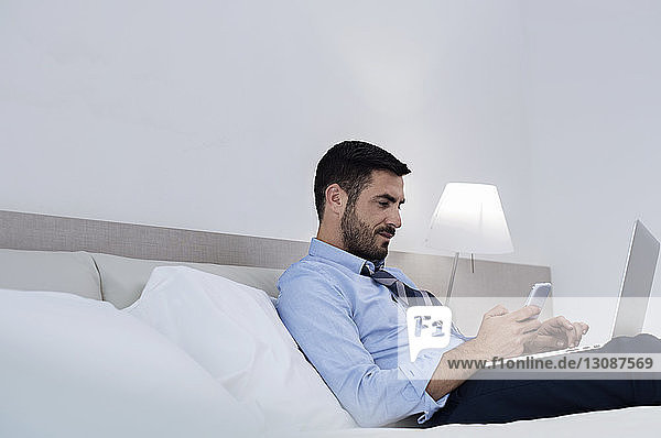 Seitenansicht eines Geschäftsmannes  der ein Smartphone und einen Laptop benutzt  während er im Hotelzimmer auf dem Bett sitzt