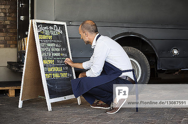 Männlicher Verkäufer in voller Länge schreibt auf Tafel  während er mit einem Speisewagen an der Straße kauert
