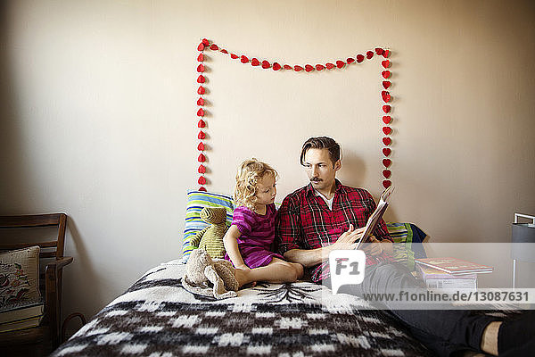 Vater zeigt der Tochter ein Buch  während er zu Hause im Bett an der Wand sitzt