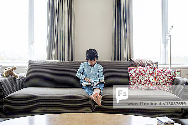 Boy reading book while sitting on sofa at home