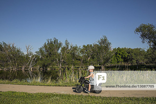 Side view of boy riding toy motorcycle by lake at park during sunny day