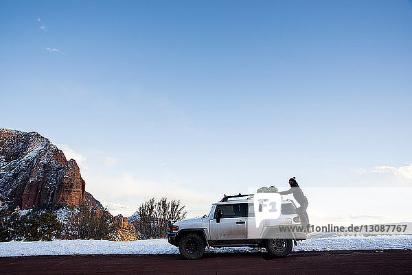 Seitenansicht eines Mannes  der im Winter in der Wüste seinen Rucksack auf einen Geländewagen gegen den Himmel stellt