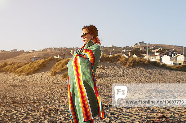 In ein Handtuch gewickelte Frau steht bei Sonnenuntergang auf Sand