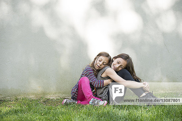 Side view of girl embracing sister while sitting against wall at yard