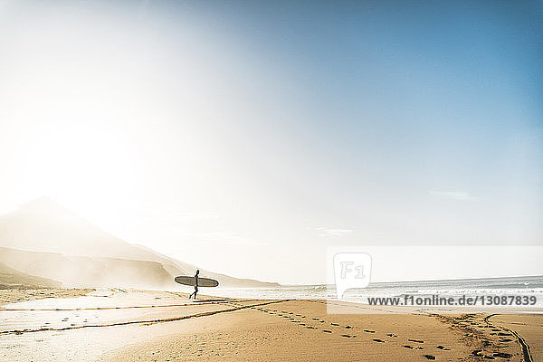 Mann mit Surfbrett geht bei Nebel am Strand gegen den Himmel in Richtung Meer