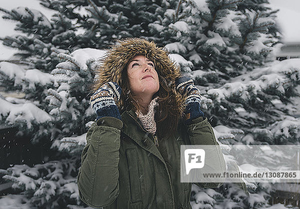 Woman looking up while wearing fur coat against trees during snowfall