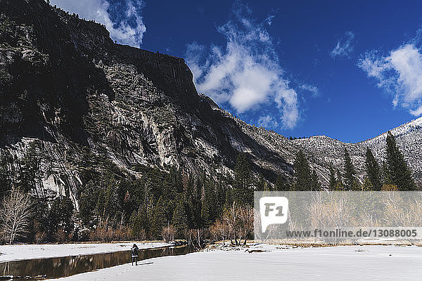 Hiker standing by river on snow against mountains at Yosemite National Park during winter