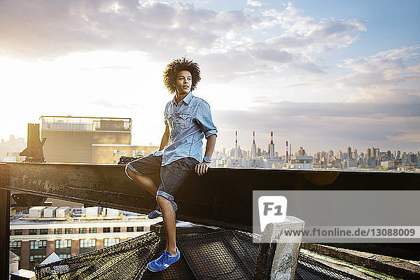 Young man sitting on metal support beam against sky during sunset