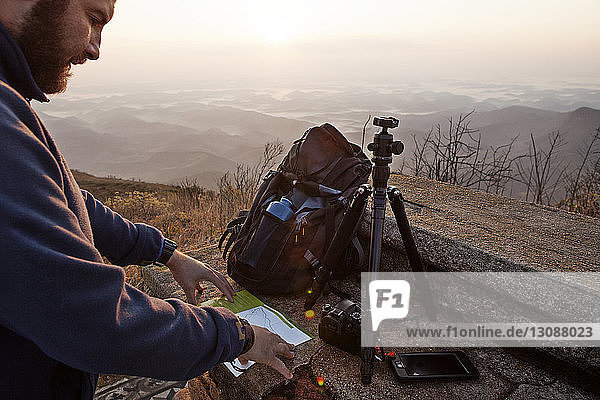 Mann liest auf der Karte  während er bei Sonnenuntergang auf einem Berg steht