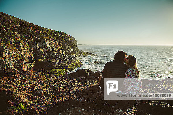 Rear view of couple kissing while sitting on rock by sea against clear sky