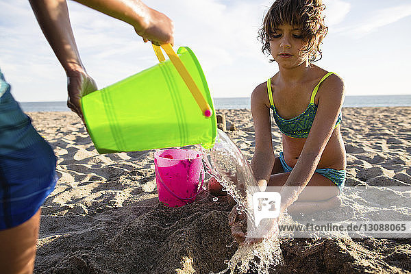 Junge gießt Wasser auf die Hände seiner Schwester  während er eine Sandburg am Strand baut