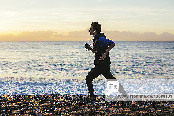 Side view of teenage boy listening music while jogging at beach against sea and cloudy sky