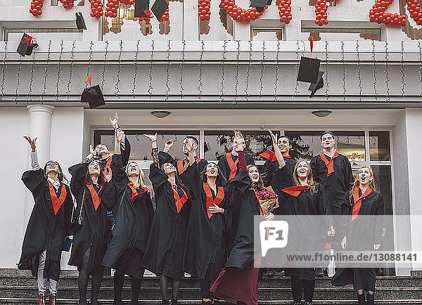 Niedriger Blickwinkel auf glückliche Studenten in Graduationskleidern  die auf Stufen am Gebäude stehen