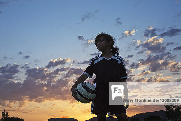 Low angle view of girl holding soccer ball against sky during sunset