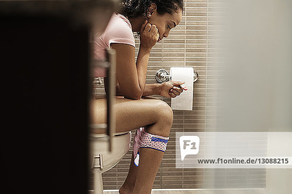 Seitenansicht einer Frau,  die einen Schwangerschaftstest macht,  während sie zu Hause im Badezimmer sitzt