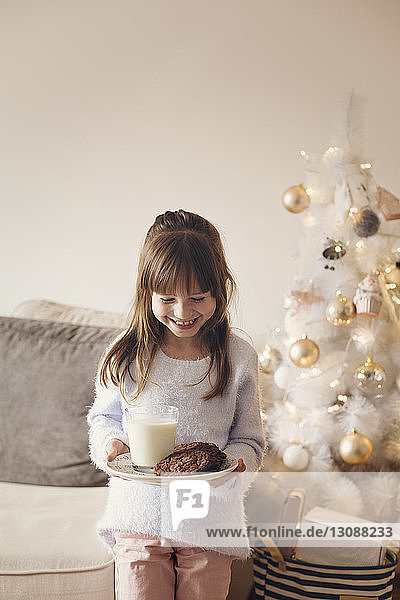 Glückliches Mädchen hält Teller mit Keksen und Milchglas  während sie zu Hause steht