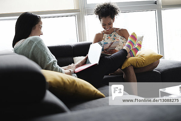 Woman using laptop computer while girlfriend looking at brochures while sitting on sofa in living room