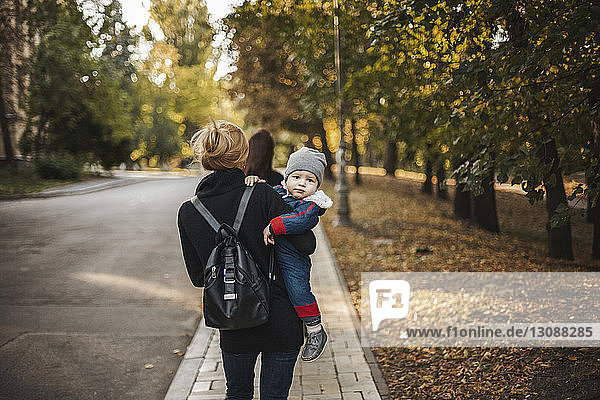 Porträt eines Sohnes  der im Herbst von der Mutter auf einem Fußweg im Park getragen wird