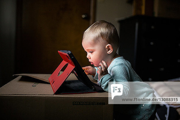 Side view of baby girl using tablet computer while standing by box at home