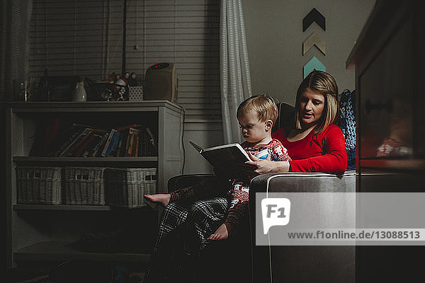 Mother with son reading book while sitting on armchair at home
