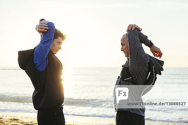 Side view of father and son stretching arms while standing face to face at beach against clear sky