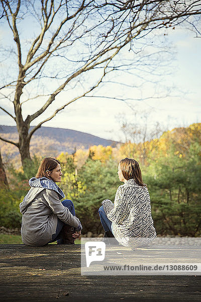 Daughter and mother talking while sitting on log against trees in forest