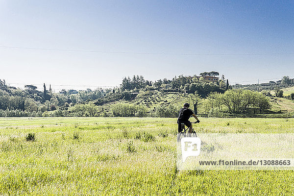 Rückansicht eines jungen Mannes beim Mountainbiken auf einem Grasfeld vor klarem Himmel an einem sonnigen Tag