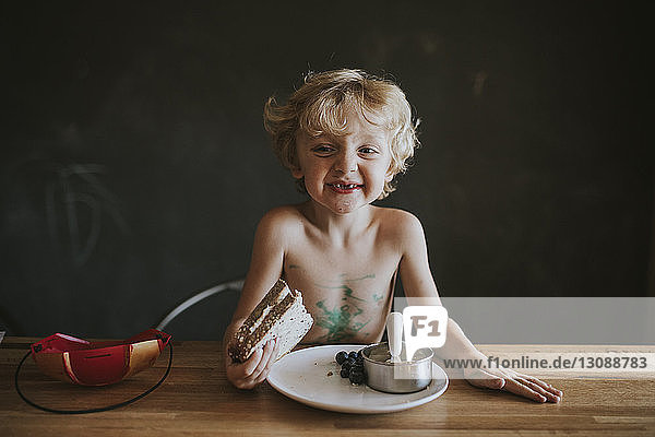 Lächelnder unordentlicher Junge hält Sandwich  während er am Tisch gegen die Tafel steht