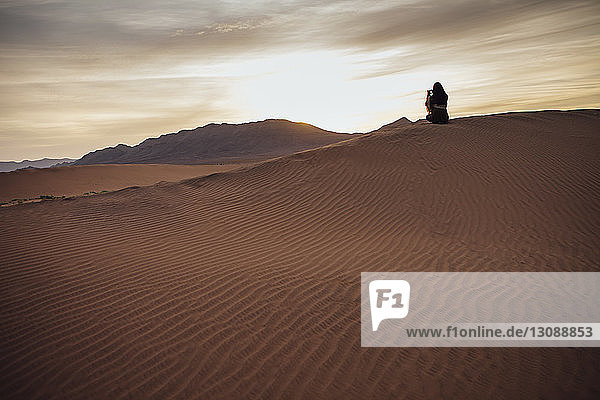 Rückansicht einer Frau  die während des Sonnenuntergangs in der Wüste per Handy fotografiert