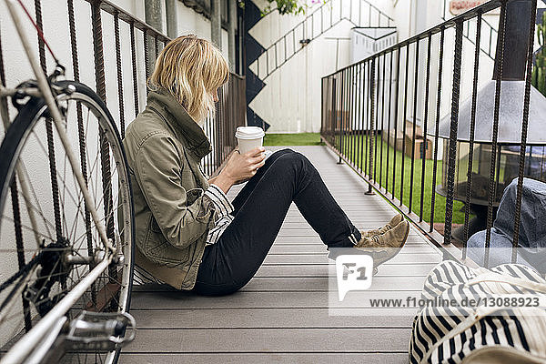 Seitenansicht einer jungen Frau  die Einwegglas hält  während sie auf einem Fußweg im Freien sitzt