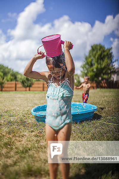 Girl pouring water on head while standing on grassy field at backyard