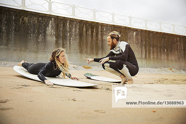 Mann spricht mit Frau  die am Strand auf einem Surfbrett liegt