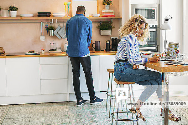 Woman using smart phone while man working at kitchen counter
