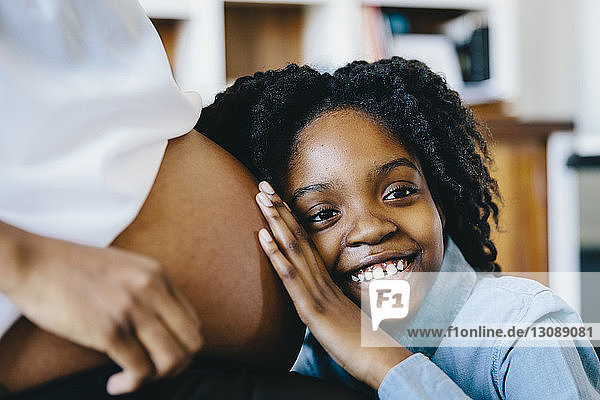 Glückliche Tochter hört zu  wie der Bauch der schwangeren Mutter zu Hause sitzt