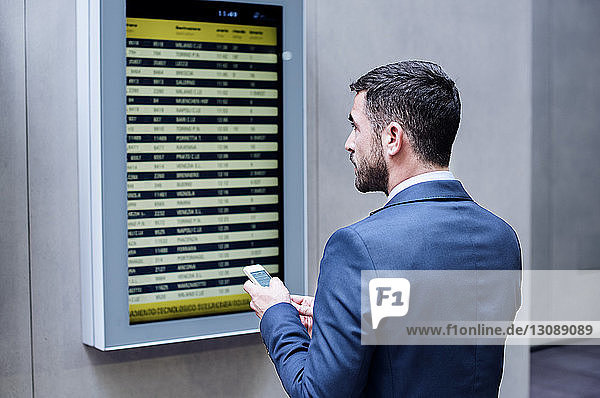 Rückansicht eines Geschäftsmannes  der ein Smartphone in der Hand hält  während er in der U-Bahn-Station die Ankunft-Abfahrtstafel liest